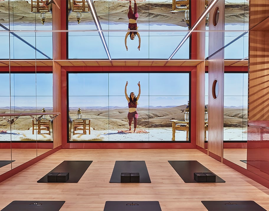 The virtual studio features wood panelled floors and floor to ceiling mirrors. Black ALO Yoga mats line the floor