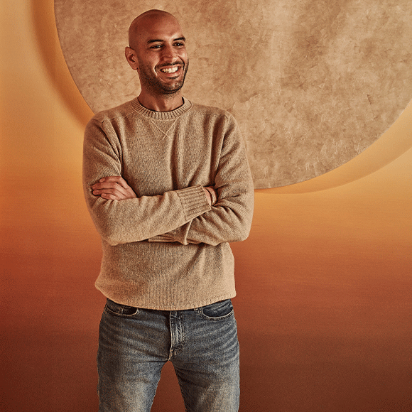 Smiling man with a shaved head wearing a beige sweater and jeans, standing with arms crossed in front of a warm-toned abstract background with a large circular shape.