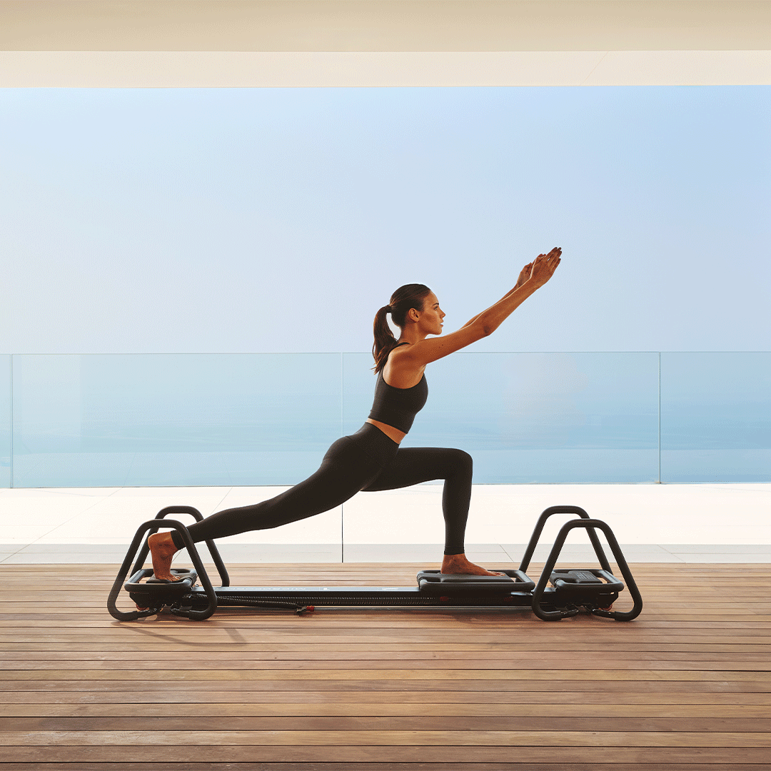 Woman in black athletic wear performing a lunge on a reformer Pilates machine on a wooden deck, with a serene ocean view and clear blue sky in the background.