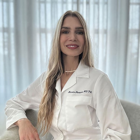 Woman in a white lab coat sitting on a chair, smiling, with embroidered name “Macrene Alexiades MD PhD” visible.