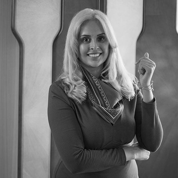 Black and white portrait of a smiling woman with long blonde hair, wearing a scarf and long-sleeved dress.