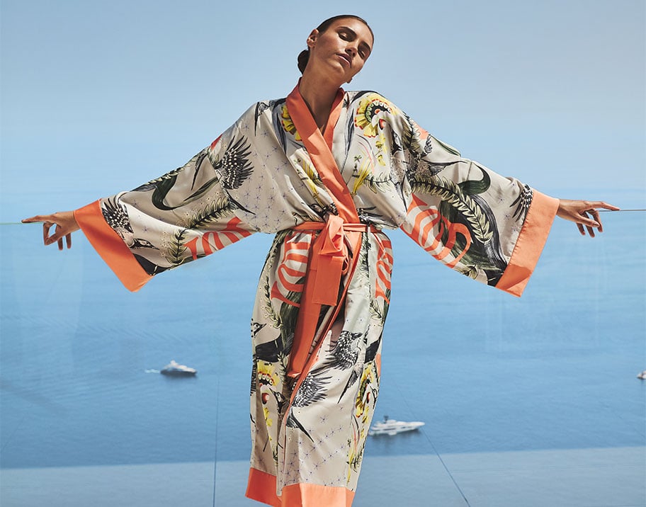 Woman in a patterned silk robe with orange trim stretches her arms out on a terrace overlooking the sea.