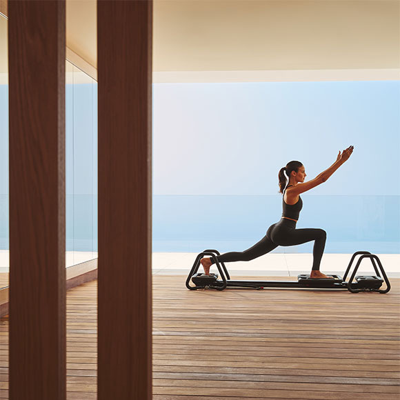 Woman performing lunging exercise on reformer machine in serene studio with panoramic sea view.