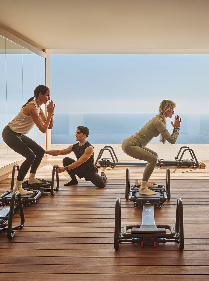 Two women practicing reformer-style exercises with an instructor in a bright studio overlooking the sea.