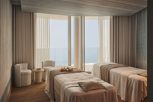 Serene spa room with two massage beds, soft lighting, ocean view through floor-to-ceiling windows, and minimal decor.