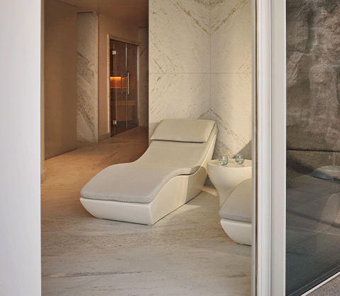 A serene marble-lined wellness space with a curved white lounger and side table, softly lit and opening onto a natural rock wall outside.